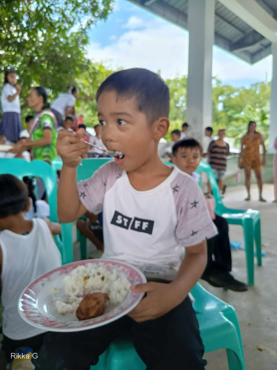 A boy holding a plate of rice and chicken, taking a big spoonful while seated in a green chair.
