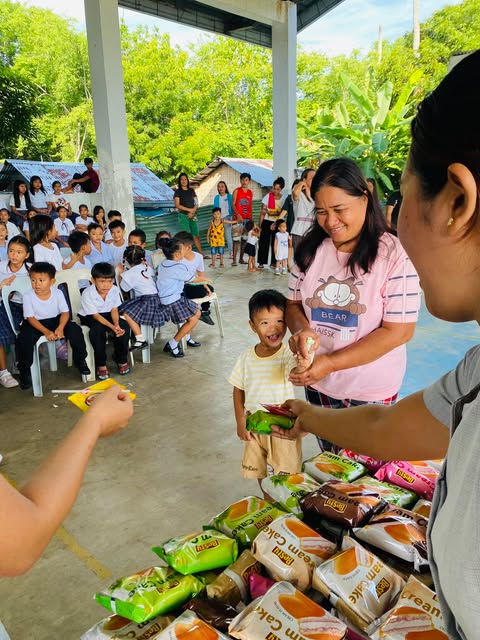 A smiling boy holding a snack next to his mother while other students wait in chairs behind them.