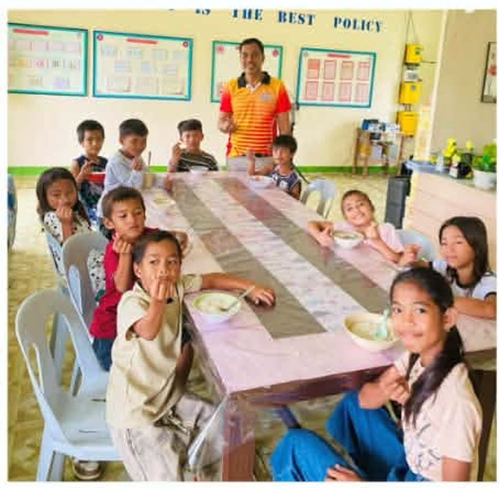Students at Naborot Carles Elementary School sharing a warm meal of rice porridge around a classroom table, with a teacher standing behind them.