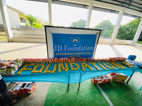 A table under the covered court at Naborot Carles Elementary School, lined with school supplies and snacks in front of a JD Foundation banner, with the word FOUNDATION spelled out in pencils.