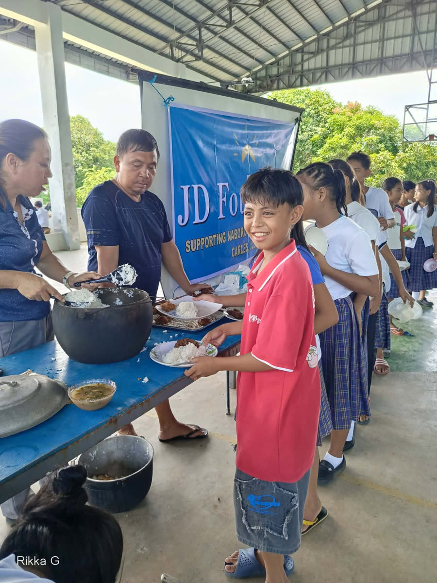 A smiling student in a red shirt receiving a plate of rice and fried chicken in front of the JD Foundation banner.