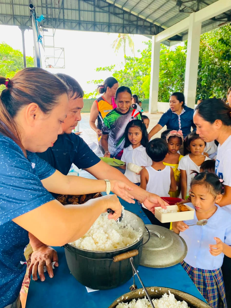 Volunteers serving rice from a large pot to students and parents lined up under the covered court.