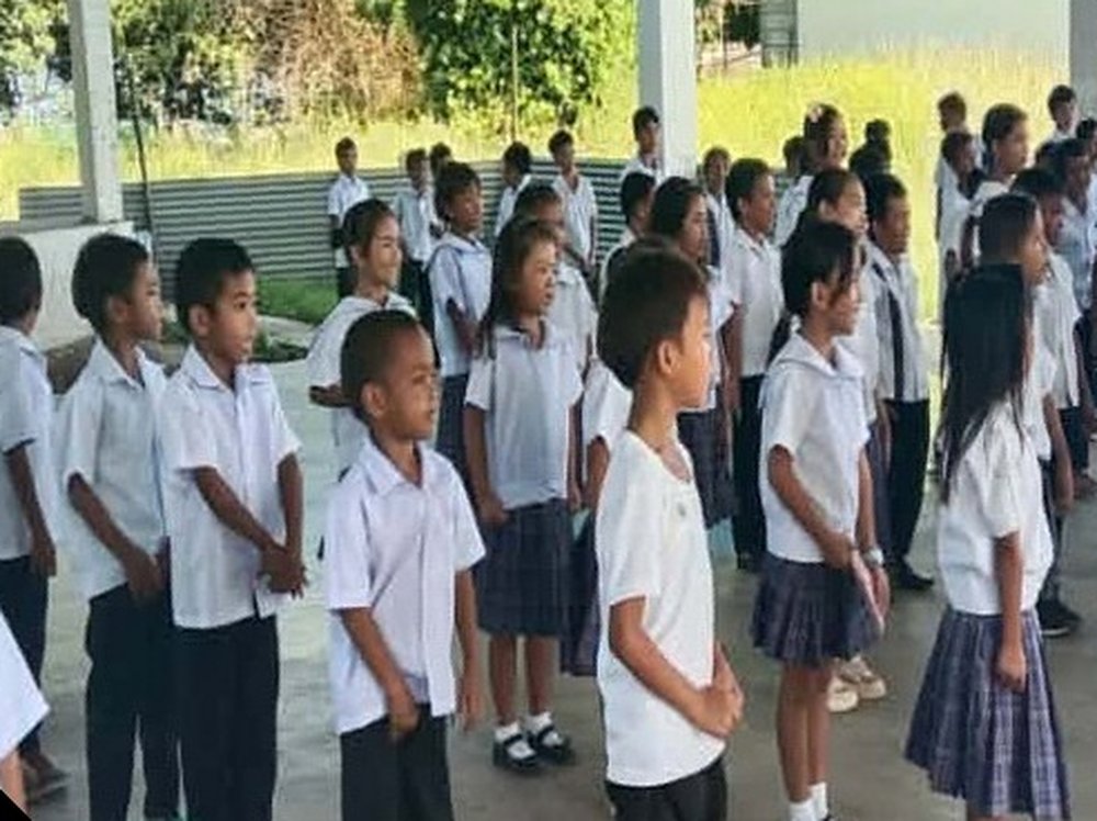Students in white uniforms standing in rows during a morning assembly under the covered court at Naborot Carles Elementary School.